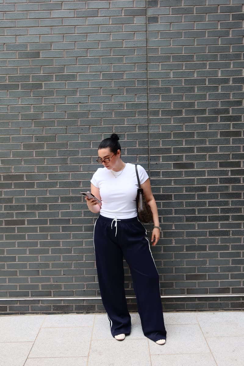 Jessie in a white tee and navy track pants against a gray brick wall, checking her phone