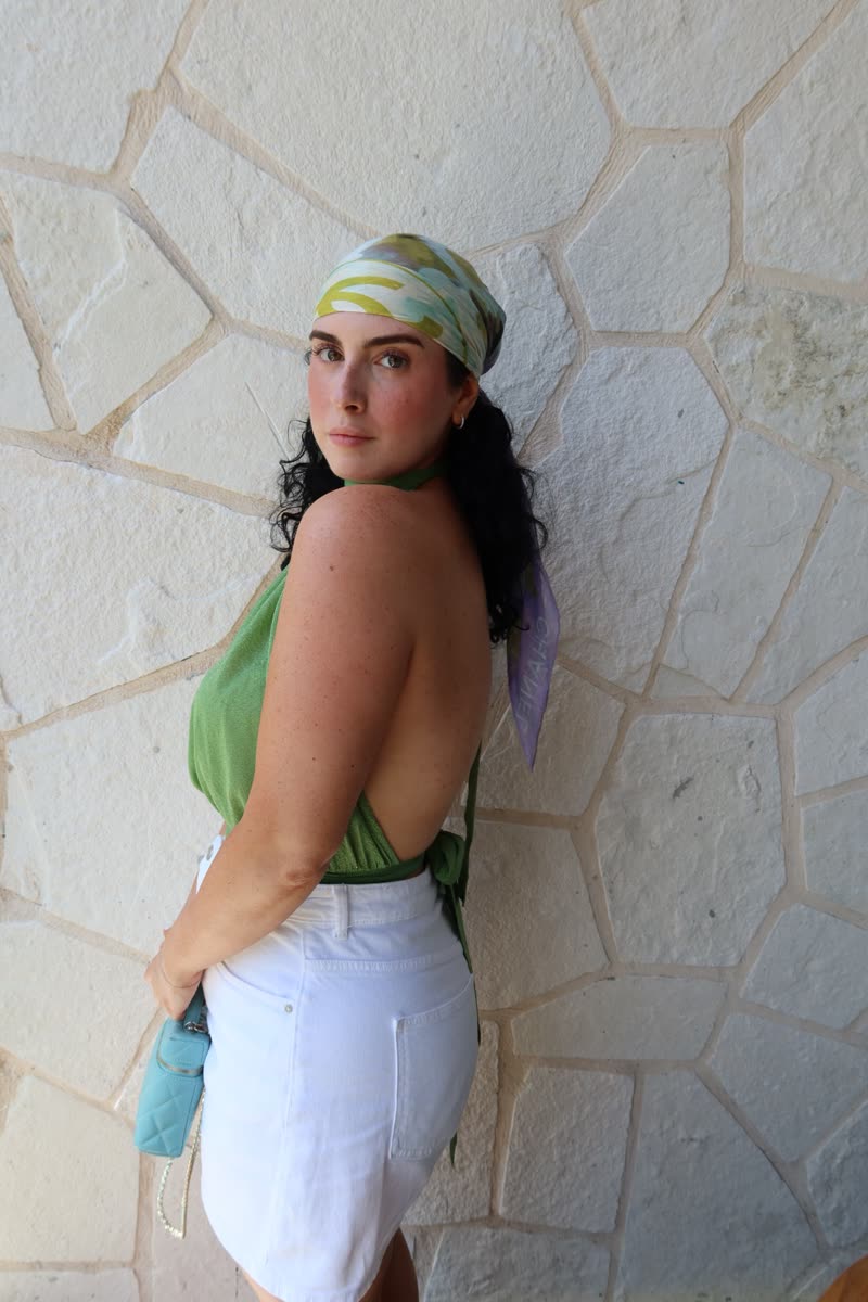 Jessie in a green halter top, white denim and a printed silk head scarf against a stone wall