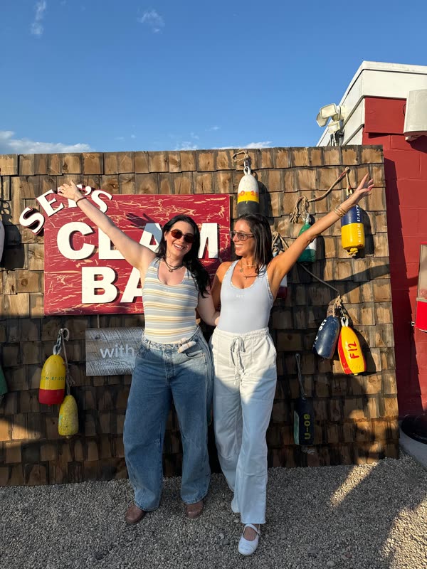 Jessie and Lindsey Carter with their arms raised in front of a clam bar sign on a sunny day